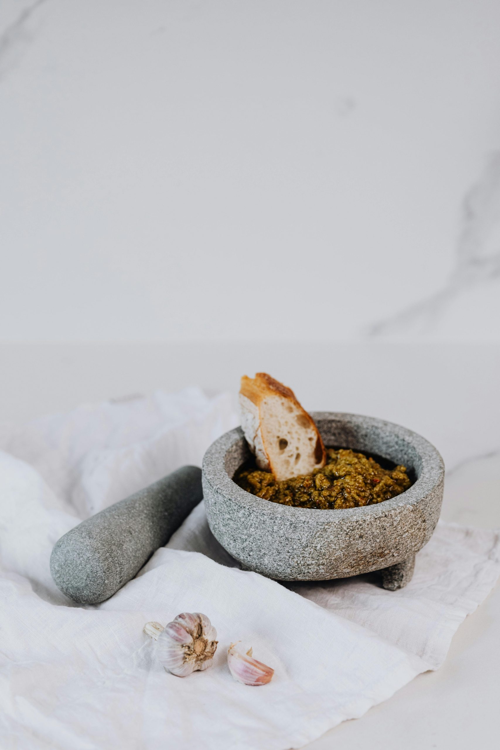 Una ciotola di pesto scuro con un pezzo di pane intinto al centro, posta su un panno bianco